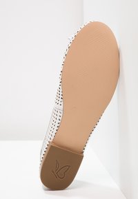 Semelle de chaussure en cuir blanc perforé avec semelle beige lisse et logo papillon sur le talon, présentée sur fond blanc.