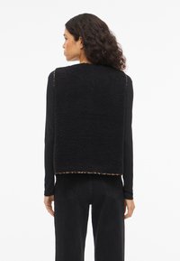 Black fleece vest with a textured finish, cropped design, and leopard print trim at the hem. Worn over a long-sleeve black shirt.