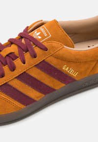 Baskets Adidas Gazelle en daim orange avec des rayures et des lacets bordeaux. Détail doré "Gazelle" au-dessus des rayures latérales. Semelle en caoutchouc.
