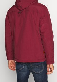 Bordeauxrote Jacke mit Kapuze, die eine glatte Textur und verstellbaren Verschluss aufweist. Kombiniert mit blauen Denim-Jeans. Klare Linien, minimale Details.