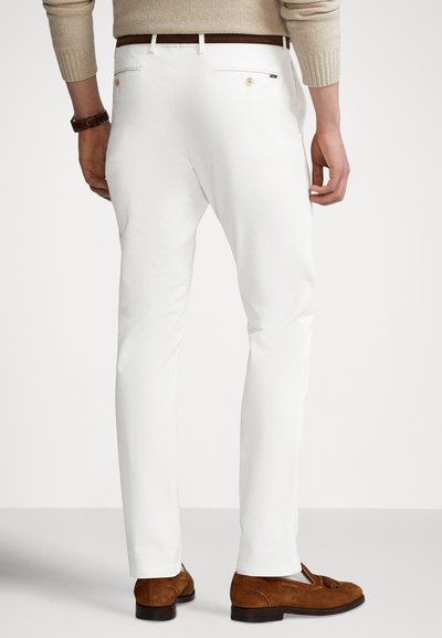 Białe bawełniane spodnie o dopasowanym kroju, z tylnymi kieszeniami i brązowymi skórzanymi detalami. Są zestawione z brązowymi zamszowymi mokasynami.