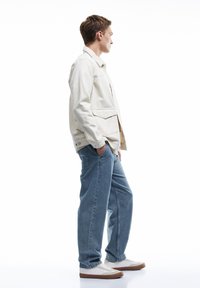 Chaqueta de lona beige clara con dos bolsillos frontales, combinada con unos vaqueros azules de corte relajado y unas zapatillas blancas con suelas marrones. Diseño simple.