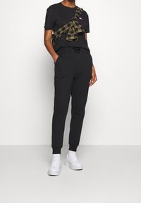 Zwarte t-shirt met een logo op de borst, gecombineerd met zwarte joggerbroek. Een groen geruit overhemd is om de t-shirt geknoopt. Witte sneakers gedragen.