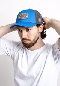 Blue baseball cap with mesh sides, featuring a gray emblem and white stitching. Worn by a person in a white t-shirt.