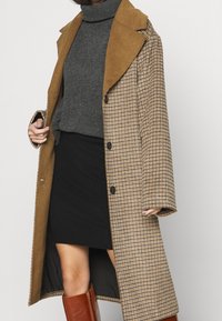 Brun rutigt jacka med en beige mocka krage, långa ärmar och svarta knappar, bärs över en grå polotröja och svart kjol.