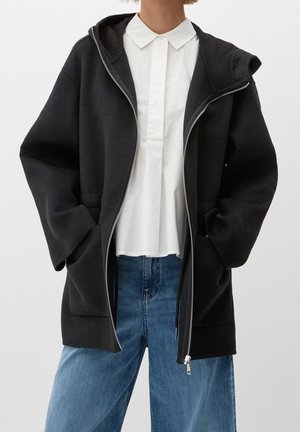 Personne portant un sweat à capuche noir déboutonné par-dessus une chemise blanche à col et un jean bleu, les mains dans les poches du sweat à capuche, cadrée au niveau du cou et des cuisses.