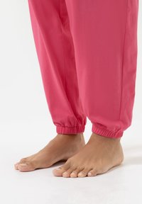 Roze elastische joggers met een gladde stofstructuur, gedragen op blote voeten, waarbij de blote voeten zichtbaar zijn en de nadruk ligt op het onderste gedeelte van de broek.