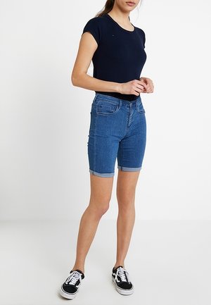 Vrouw draagt een donkerblauw kort mouwtopje, mid-lange opgerolde spijkershorts en zwart-witte sneakers, staand met handen in elkaar.