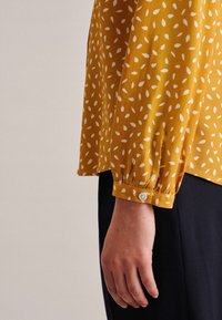 Mustard blouse featuring a leaf pattern in white, with long sleeves gathered at the cuffs and a button detail at the wrist.