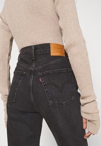 Jeans de mezclilla negros de cintura alta con un parche trasero de cuero marrón claro, que cuentan con dos bolsillos traseros y un ligero desvanecimiento en el color.