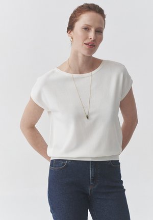 Bílá blůza s krátkým rukávem, kulatým výstřihem, hladkým materiálem a uvolněným střihem, doplněná tmavě modrými džínami a zlatým náhrdelníkem.