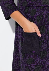 Robe noire avec un motif abstrait enroulé violet, dotée d'une poche violet uni accentuée par de petits points et un logo.