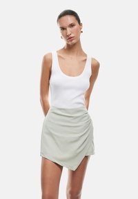 Vrouw met strak naar achteren gekamd haar, draagt gouden oorbellen, een witte mouwloze ribbeltop en een lichtgrijze asymmetrische wikkelrok.