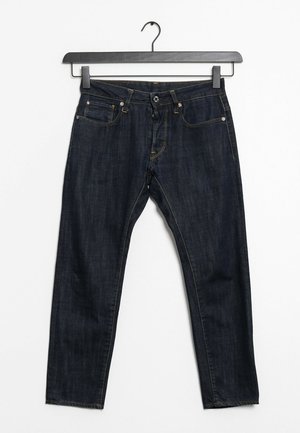 Tmavě modré džíny z denimu visící na černém ramínku na bílém pozadí, zobrazující přední kapsy, knoflík a detaily zipu.