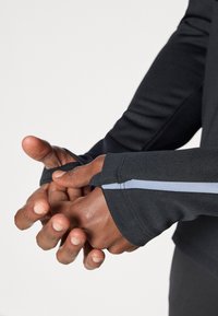 Must mustaga shirt, mille pikkade varrukatega, soonikute ja helesinise aktsendiga randmel. Sile kangas ja keha külge kloobiv disain. Käed on ees ristis.