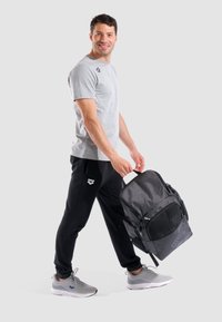 Maglietta sportiva grigia e pantaloni della tuta neri. L'uomo tiene uno zaino grande nero con dettagli in rete e cerniere. Le scarpe sono sneakers grigie.
