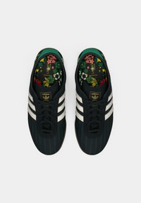 Αθλητικά παπούτσια Adidas με μαύρο σχεδιασμό ρίγες, τρεις λευκές ρίγες, μαύρα κορδόνια και πολύχρωμο εσωτερικό πέλμα με λουλουδάτο μοτίβο.