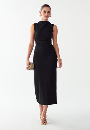 Femme portant une robe midi noire sans manches, des boucles d'oreilles dorées, un bracelet, tenant une pochette beige et des sandales dorées à talons hauts et à brides sur fond blanc.