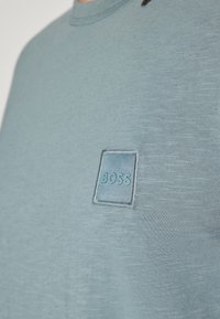 Gros plan d'un t-shirt bleu clair à col rond avec une petite étiquette rectangulaire portant l'inscription "BOSS" sur la poitrine.