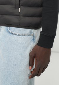 Chaleco acolchado negro con diseño de acolchado, combinado con una camiseta de manga larga oscura y jeans de color azul claro, que muestra una textura de tela suave.