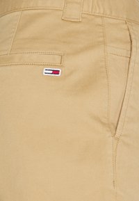 Beżowe spodnie z tylną kieszenią typu welt i małą prostokątną naszywką w kolorach białym, czerwonym i granatowym, wyszywaną nad kieszenią.