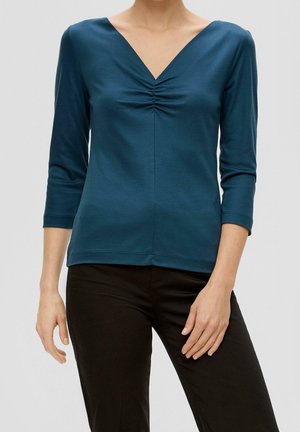 Kobieta w turkusowej bluzce z długim rękawem o dekolcie w serek z marszczonym detalem na środku i czarnych spodniach, stojąca na jednolitym jasnym tle.