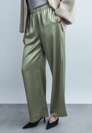 Femme portant un pantalon ample en satin vert olive, une veste beige, un haut gris et des escarpins noirs à bout pointu, debout devant un fond gris clair.