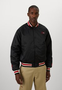 Black bomber jacket with red and white striped ribbed collar and cuffs, featuring a front button closure and logo detail on the chest.