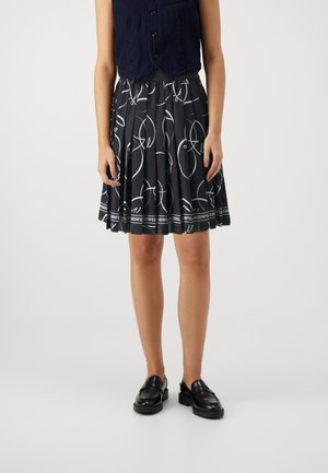 Personne portant une veste sans manches bleu marine, une jupe noire à motif abstrait blanc avec bordure à logo, et des mocassins noirs, debout devant un fond uni.