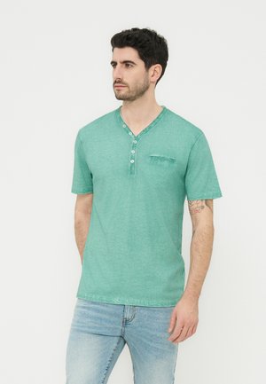 Homme aux cheveux foncés et à la barbe, portant un t-shirt henley à manches courtes vert et un jean bleu clair, debout devant un fond uni.