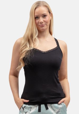 Femme aux longs cheveux blonds portant un débardeur noir bordé de dentelle et un pantalon bleu clair avec un imprimé grue, mains dans les poches, fond neutre.