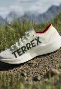 Λευκό παπούτσι τρεξίματος Terrex με κίτρινα κορδόνια που ακουμπά σε χαλίκι, με γρασίδι στο φόντο και θολά βουνά στο βάθος.