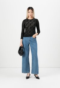 Svart blonde langermet topp med blomsterdesign, parret med lyseblå høyt liv vide bukser. Holder en svart håndveske og har på seg spisse leiligheter.