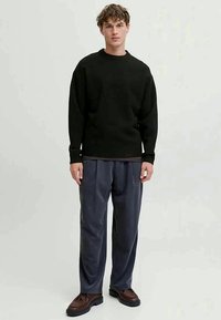 Young man standing wearing black sweater, loose dark gray trousers, and brown leather shoes on light background.