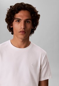 Junger Mann mit lockigem dunklem Haar, der ein schlichtes weißes Calvin Klein T-Shirt trägt und mit neutralem Gesichtsausdruck direkt in die Kamera schaut.