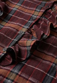 Gros plan sur un tissu à carreaux rouge, noir et marron avec des boutons noirs et un détail de couture froncée le long de la ligne des boutons.