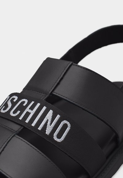 Sandale noire avec larges brides en cuir et une bande en tissu noir ornée du texte "MOSCHINO" brodé en blanc.