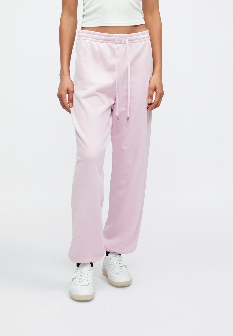 Ροζ ανοιχτόχρωμο παντελόνι φόρμας από απαλό ύφασμα, με ελαστική μέση και κορδόνια, εφαρμογή σε χαλαρή γραμμή, συνδυασμένο με λευκά αθλητικά παπούτσια.