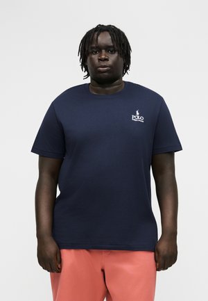 Άνδρας με μεσαίου μήκους dreadlocks φοράει μπλε ναυτικό μπλουζάκι Polo Ralph Lauren και κοραλλί σορτς, στέκεται μπροστά σε απλό ανοιχτό φόντο.