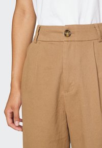 Pantalon beige tailored avec une texture lisse, taille haute et une fermeture à un bouton. Présente des plis avant structurés et des poches latérales.