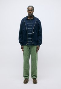 Felpa con zip blu sopra una maglietta a righe blu e bianche, abbinata a pantaloni larghi verdi e scarpe marroni. Sfondo semplice.