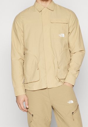 Mężczyzna ubrany w beżową, długorękawową kurtkę i dopasowane spodnie z wieloma kieszeniami oraz logo The North Face na klatce piersiowej i udzie.