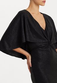Svart klänning med djup V-ringning, flödande capeärmar, strukturerad tyg med en subtil lyster och en knutdetalj i midjan.