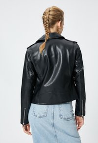 Svart läderbikerjacka med en blank finish, dragkedjeslutningar och strukturerade axlar, bärs över en ljusblå denimkjol.