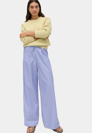 Donna in piedi con le braccia incrociate, indossa un maglione giallo chiaro, pantaloni a gamba larga a righe blu e bianche, e sandali neri.