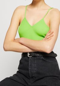 Bez rękawów, prążkowany top na ramiączkach w jaskrawej zieleni. Dekolt w serek z cienkimi ramiączkami. Noszony z wysokim stanem czarnych dżinsów i czarnym paskiem.