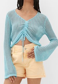 Top de punto ligero y translúcido en azul claro con escote en V, detalle fruncido en la parte delantera, y mangas largas acampanadas, combinado con pantalones cortos de mezclilla amarillos.