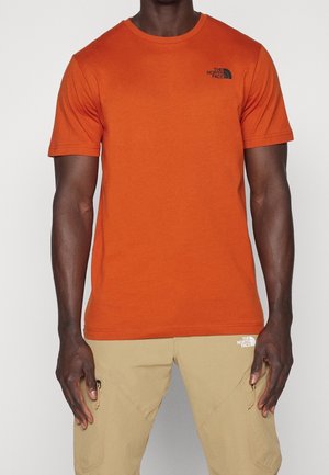 Mann trägt ein orangefarbenes North Face T-Shirt mit kurzen Ärmeln und beige North Face Hose mit Reißverschlusstaschen, steht vor einem einfarbigen Hintergrund.