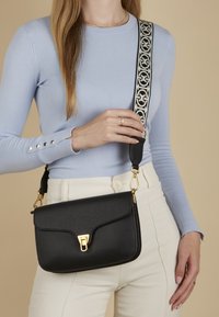 Sac bandoulière en cuir noir avec une finition texturée, quincaillerie dorée et une sangle ajustable à motifs. Le modèle porte un haut bleu clair.