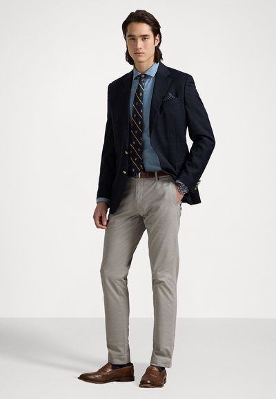 Blazer azul-marinho sobre uma camisa de denim clara, emparelhado com uma gravata riscada. Chinos bege e sapatos loafers castanhos completam o visual. Acessórios notáveis incluem um lenço de bolso.